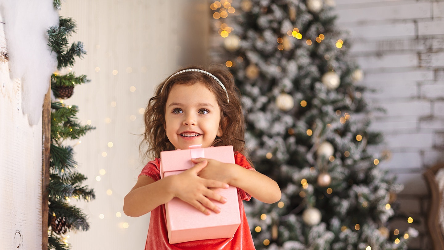 A little girls holding a pink Christmas present in front of a Christmas tree for a guide to Christmas for separated parents