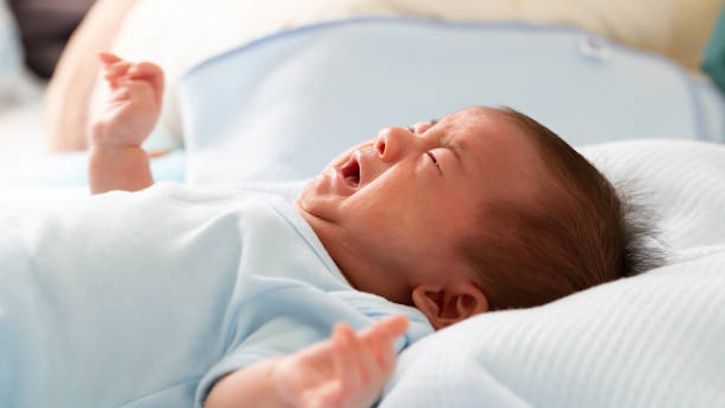 A newborn baby dressed in blue, lying a bed and crying