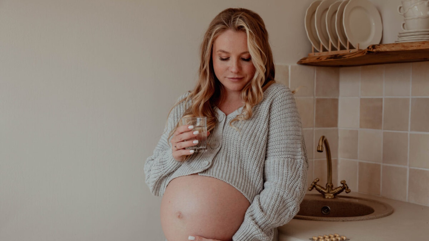Pregnant and aspirin: a pregnant woman drinking a glass of water with a pack of pills on the kitchen counter